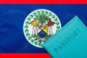 A teal passport partially covers the national flag of Belize, inviting travelers to explore its charm. The flag showcases a coat of arms with two figures, tools, and a shield surrounded by green leaves on a white disk, all set against a blue background with red stripes at the top and bottom.