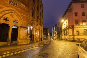 A quiet, dimly lit urban street in Manchester at night offers a traveler unmissable experiences. Historic brick and stone buildings stand illuminated by streetlights, with wet pavement hinting at recent rain. Under a deep blue, cloudy sky, the scene invites exploration without pedestrians or vehicles in sight.